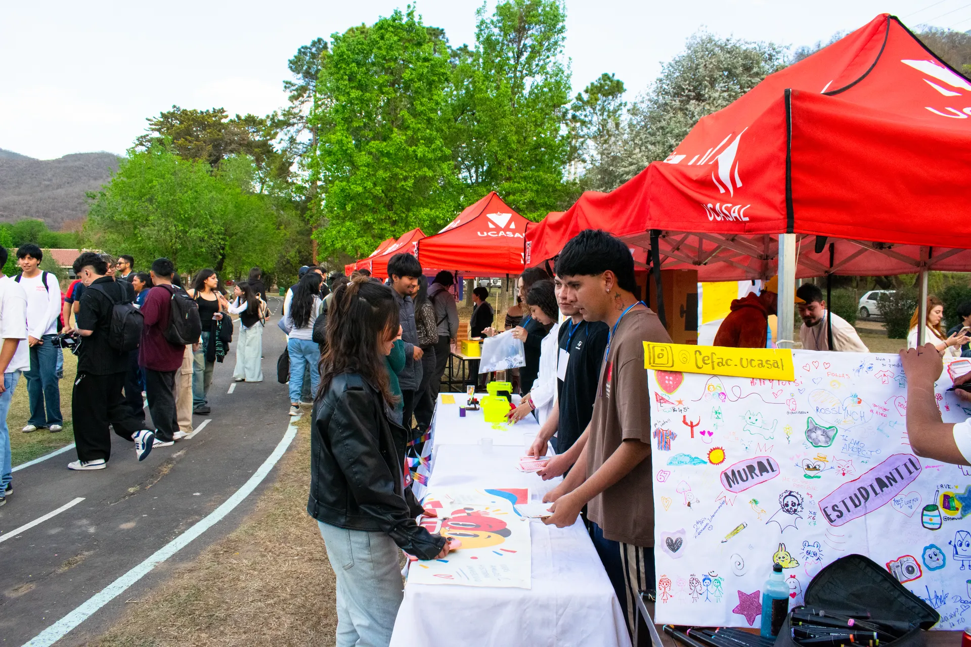 Fila de carpas rojas junto al camino del campus; estudiantes pintan un mural sobre una mesa.
