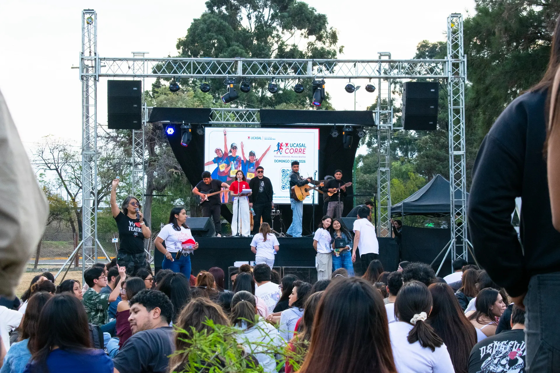 Una banda toca en el escenario con pantalla mientras el público mira sentado en el césped.