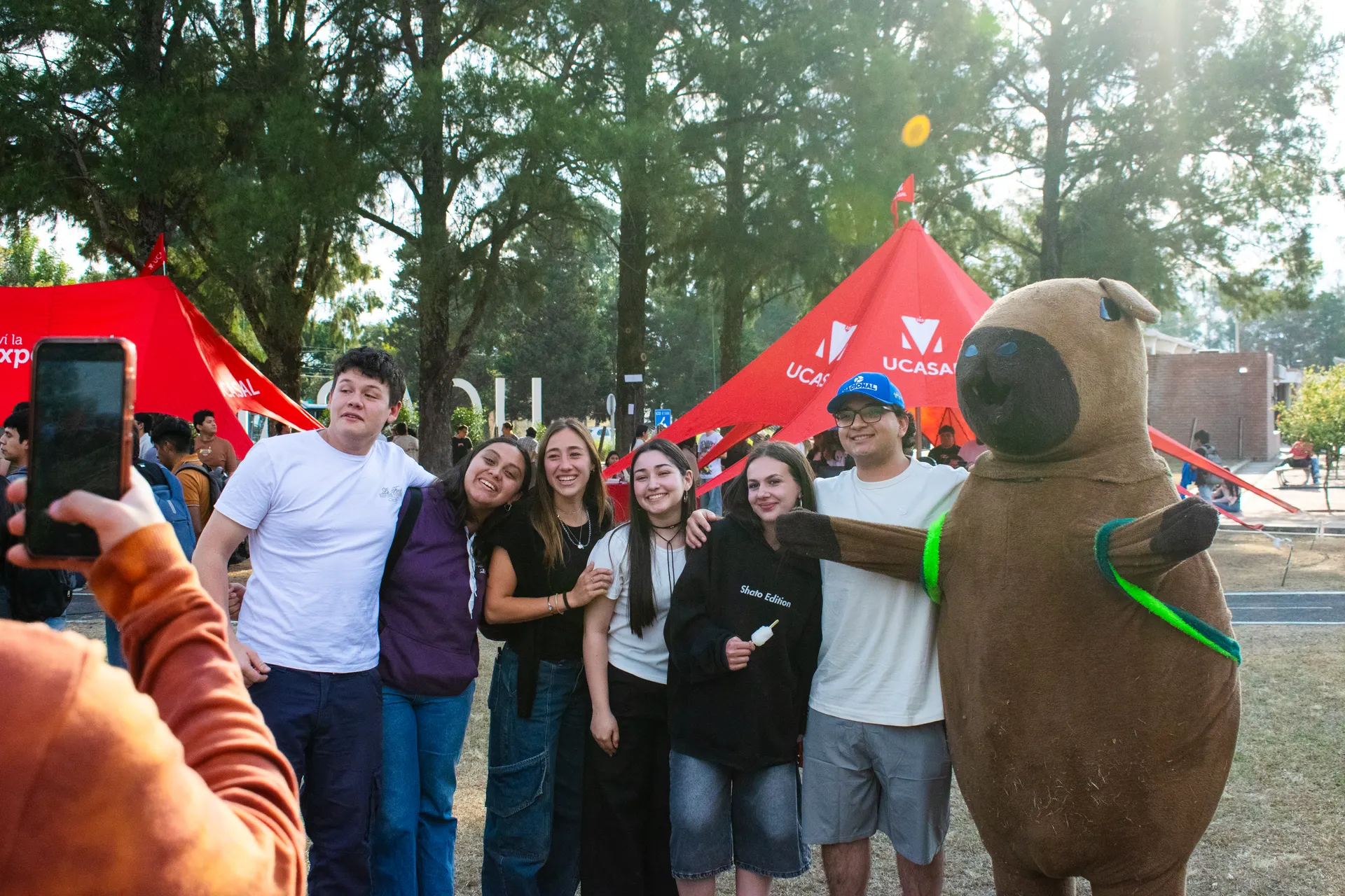 Grupo de estudiantes posa con el carpincho del Centro bajo las carpas rojas.