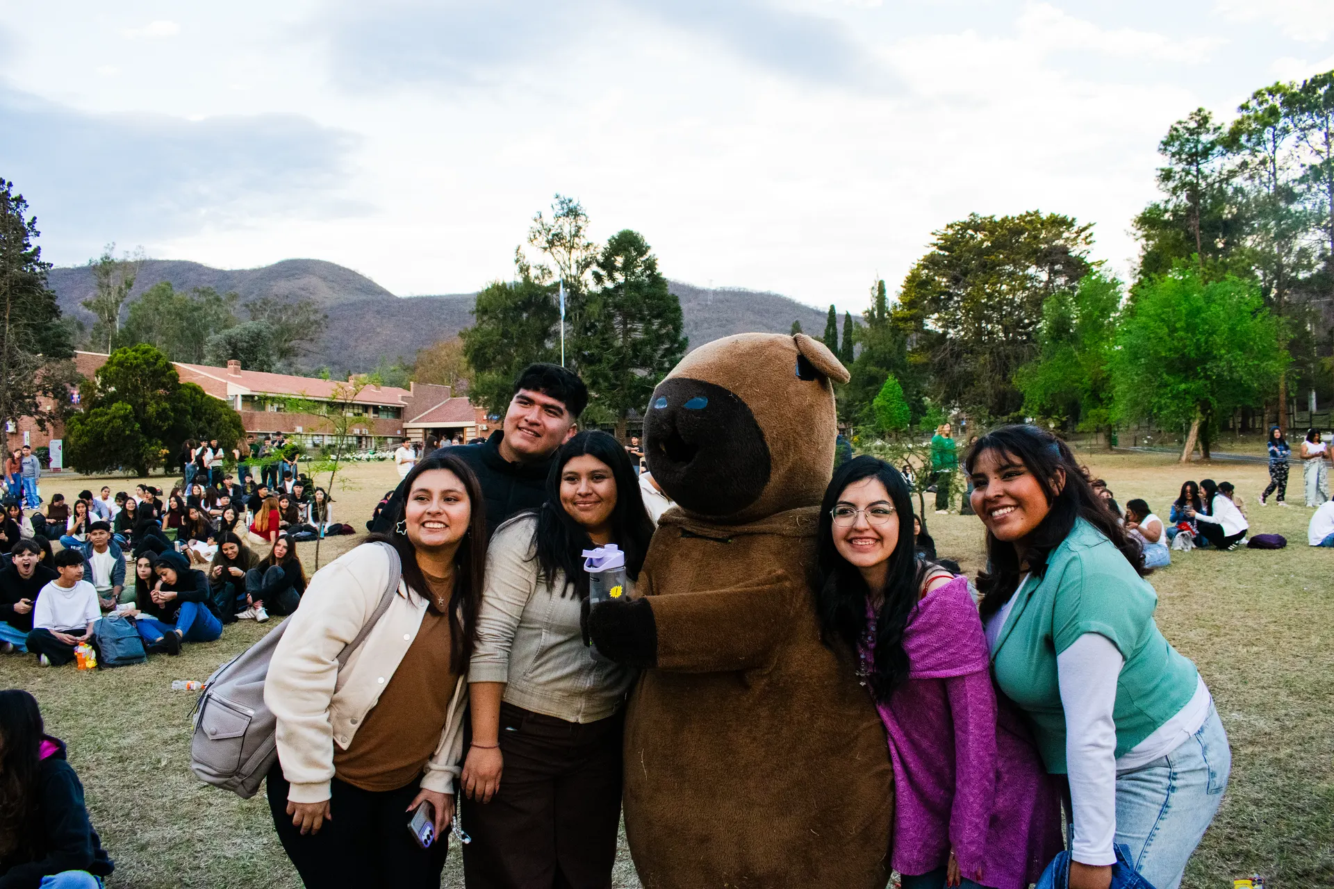 Grupo de estudiantes abrazados con el carpincho del Centro en el césped, con los cerros de fondo.